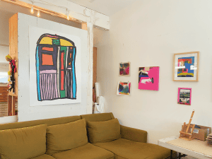 A large, colorful artwork filled with bold lines and abstract shapes sits on a wall. Below it, a comfy looking couch. On the adjacent wall sit five smaller artworks of varying size.