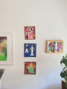 In a vertical line, three small paintings on cardboard sit on a wall. Each depict colorful, costumed, faceless figures. To the right and left are more gestural and abstract framed works. A potted plant peeks into the left side of the image.